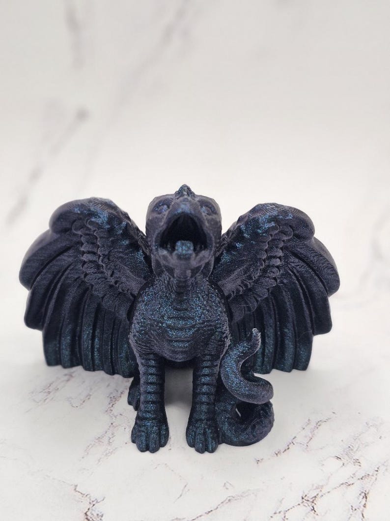 May include: A dark blue griffin figurine with outstretched wings and an open beak. The mythical creature has a textured surface and a curled tail. The figurine is standing on a white surface.