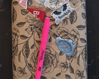 May include: A rectangular package wrapped in tan paper with a black floral print, featuring a bright pink Sharpie highlighter, a light blue wax seal, and various stickers including text reading "Bookish Baddie" and "Fell in love on the third".