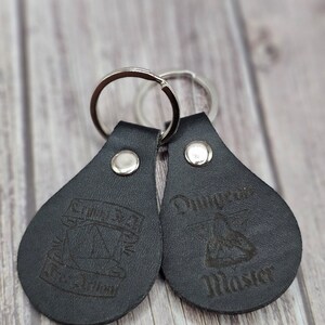 May include: Two black leather keychains with silver key rings. One keychain has the text "Save the Music" and a musical note design. The other keychain has the text "Dungeon Master" and a dice design.