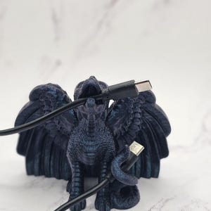 May include: A dark blue griffin figurine with outstretched wings, holding a black USB cable. The griffin has detailed feathers and a textured surface. The background is a blurred white marble.