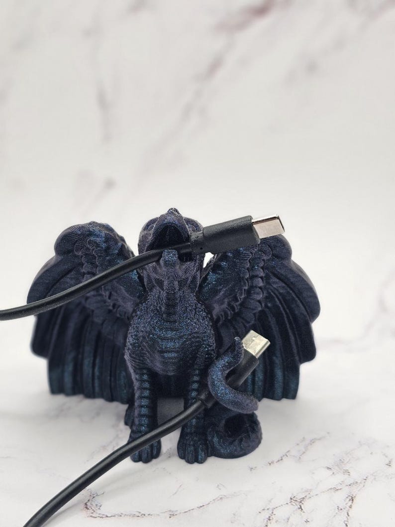 May include: A dark blue griffin figurine with outstretched wings, holding a black USB cable. The griffin has detailed feathers and a textured surface. The background is a blurred white marble.
