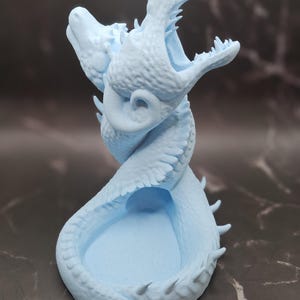 May include: A light blue 3D printed dragon head dice holder with an open mouth and a small, round, open space inside for dice.
