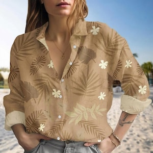 Puede incluir: Camisa abotonada color beige con un estampado de hojas y flores tropicales. La camisa de manga larga presenta cuello y puños beige, y botones blancos. El diseño incluye hojas marrones y blanquecinas y flores blancas.