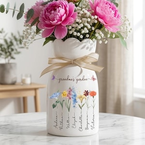 May include: A white ceramic vase with a scalloped top, tied with a beige ribbon, and filled with pink peonies and white baby's breath. The vase is decorated with floral illustrations and the text "grandma's garden".