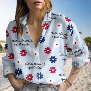 May include: Light blue button-down shirt with a floral pattern in red, white, and blue. The shirt features the text "stars, stripes, & equal rights" in black script. The shirt has long sleeves and a collared neckline.