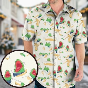 May include: Short-sleeved button-down shirt with a repeating pattern of Kermit the Frog in a yellow blazer and blue tie. The shirt is off-white with a green collar and cuffs. The background is blurred, suggesting an outdoor setting.