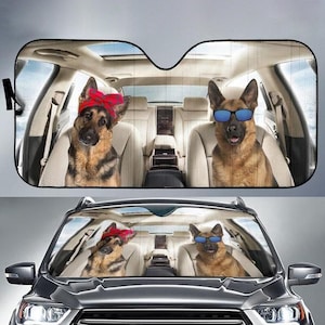 Op de afbeelding: Een zonnescherm voor de auto met twee Duitse herders die een zonnebril dragen. Een hond draagt een rode bandana. De honden zitten op de achterbank van een auto.