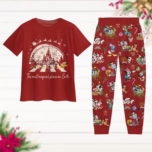 May include: Red pajama set comprising a t-shirt and trousers. The shirt displays Disney characters crossing a street in front of a castle, with the text "The most magical place on Earth." The trousers feature a repeating pattern of Disney characters in festive scenes.