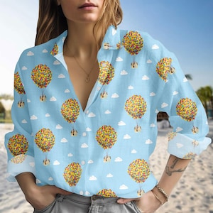 May include: Light blue button-down shirt featuring a repeating pattern of colorful hot air balloons with attached houses and white clouds. The long-sleeved shirt has a classic collar and is made of a lightweight fabric.