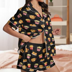 May include: Black satin pajama set featuring a repeating pattern of cartoon french fries and chicken nuggets. The short-sleeved top and shorts have white trim. The fries wear sunglasses and are in red and white striped containers.