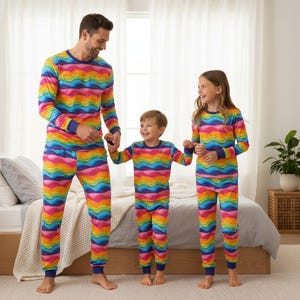 Könnte beinhalten: Drei Personen tragen passende, regenbogenfarbene Pyjama-Sets. Die Pyjamas haben lange Ärmel und Hosen mit einem wellenförmigen Muster in Rot-, Orange-, Gelb-, Grün-, Blau- und Lilatönen. Die Bündchen und Ausschnitte sind marineblau.
