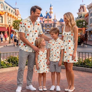May include: A family wearing matching cream-colored clothing featuring a repeating Halloween pattern of white ghosts, orange pumpkins, and black cats. The clothing includes a short-sleeved button-down shirt, a sleeveless dress, and a sleeveless child's dress.