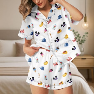 May include: White satin pyjama set with a playful pattern of cartoon icons, including ice cream cones, hats, and bows. The set features a button-down shirt with short sleeves and matching shorts, trimmed with white piping.
