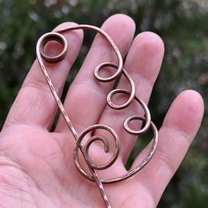 Puede incluir: Horquilla de cobre hecha a mano con un diseño abstracto único. La horquilla presenta una serie de formas en espiral y una textura martillada, creando un accesorio artístico y elegante. El cobre tiene un tono marrón rojizo cálido.