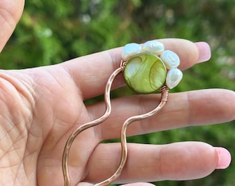 May include: A copper hair stick with a wavy design. The stick is topped with a green and white shell cabochon and a cluster of white pearls.