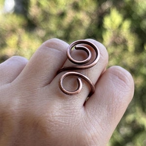 Op de afbeelding: Een koperkleurige ring met een spiraalvormig ontwerp. De ring wikkelt zich om de vinger met een wervelend patroon, waardoor een unieke en artistieke uitstraling ontstaat. Het metaal heeft een warme, roodbruine tint en het ontwerp is eenvoudig maar opvallend.
