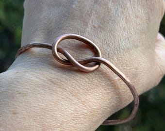 Peut inclure: Un bracelet de couleur cuivre avec un motif de nœud. Le bracelet est fabriqué à partir d'une seule pièce de métal pliée en une boucle continue, le nœud étant le point central. Un bijou simple mais élégant.