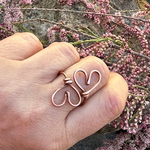 May include: A copper wire ring with two intertwined heart shapes. The ring is worn on a finger.