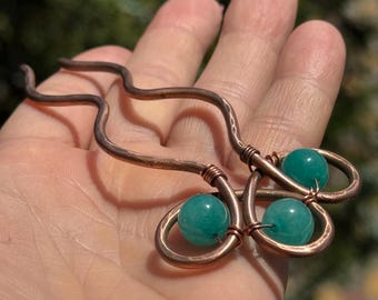May include: Copper hair pin with three teal-colored beads. The pin has a wavy design with a hammered texture. The beads are round and add a pop of color to the copper hair accessory.