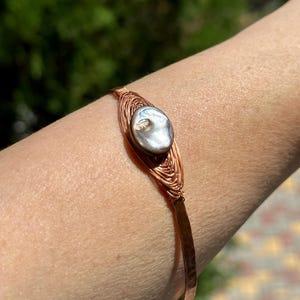 May include: A copper wire wrapped bracelet with a large, iridescent pearl in the center.