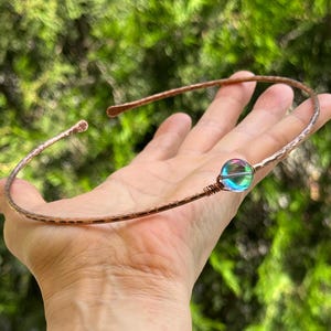May include: A copper-toned metal torque necklace featuring a hammered texture along the band. The open-ended design includes a single iridescent, multi-colored glass bead secured with wire wrapping on one side of the circular frame.