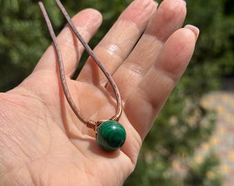 May include: A handcrafted necklace featuring a vibrant green malachite bead pendant. The pendant is suspended from a copper wire frame with a delicate, textured finish. The necklace is held in a hand against a blurred green background.