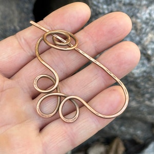 May include: A copper wire hair pin with a spiral design at the top and a curved, looped design at the bottom.
