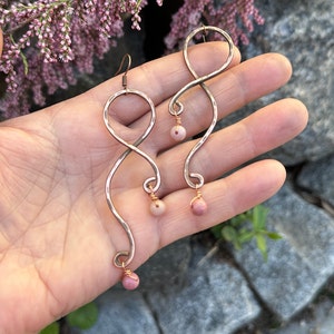 Könnte beinhalten: Handgefertigte kupferfarbene Ohrringe mit Unendlichkeitszeichen-Design. Jeder Ohrring hat eine gehämmerten Textur und einen kleinen, runden, rosa Edelstein am unteren Ende. Die Ohrringe werden vor einem natürlichen Hintergrund mit rosa Blüten präsentiert.
