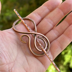 May include: A copper hair stick with a decorative treble clef design. The stick is hammered and has a rustic finish.