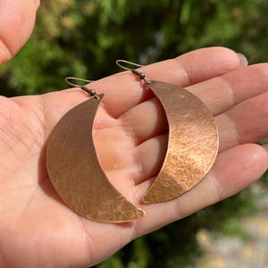 Può includere: Un paio di orecchini a forma di mezzaluna in rame con una texture martellata. Gli orecchini sono appesi alla mano di una persona.