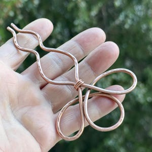Puede incluir: Un accesorio para el cabello de cobre hecho a mano con un diseño floral. El alambre de cobre tiene forma de bucles y curvas, con una textura martillada. El accesorio se sostiene en una mano sobre un fondo verde borroso.