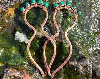 May include: Two copper hair pins with green malachite beads. The pins are twisted and have a wavy design. They are lying on a mossy rock.