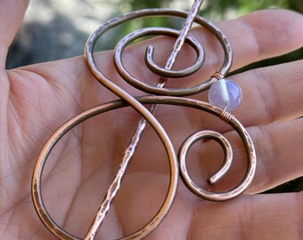 May include: Copper-toned hair pin with a unique, swirling design. The pin features a hammered texture and a small, light purple bead. The design is reminiscent of musical notes, adding a touch of artistic flair to any hairstyle.