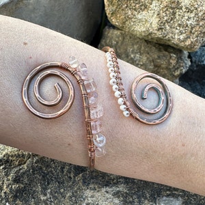 May include: A copper wire bracelet with two spirals and a row of clear quartz crystals. The bracelet is on a person's arm.