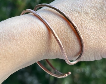 Peut inclure: Deux bracelets en cuivre martelé, de couleur brun rougeâtre, avec une surface texturée. Le design est simple, avec une forme incurvée. Les bracelets sont présentés sur le bras d'une personne. Ils mesurent environ 15-18 cm de circonférence.