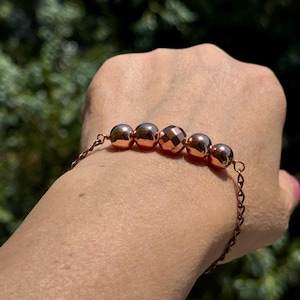 May include: A copper-colored bracelet featuring five round beads and a delicate chain. The beads have a faceted design, reflecting light. The bracelet is displayed against a blurred green background.