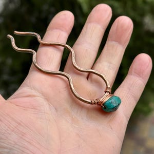 Puede incluir: Una horquilla para el pelo de cobre con un diseño ondulado y una piedra turquesa en el extremo.