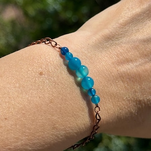 May include: A handmade bracelet featuring a series of graduated, round, light blue beads. The beads are strung on a delicate copper-colored chain. The bracelet is designed to be worn on the wrist.