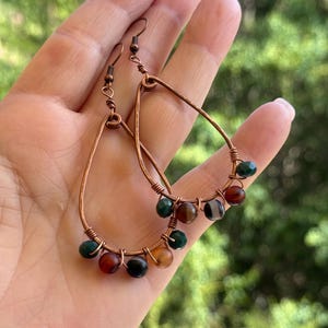 May include: Copper teardrop earrings with wire-wrapped detailing. Each earring features a series of colourful gemstone beads in shades of green, red, brown, and black. The earrings are designed with a hook closure.