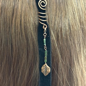 May include: A black velvet hair accessory featuring a coiled copper wire accent at the top. A delicate chain hangs from the wire, decorated with small green beads and a metallic leaf-shaped charm at the bottom, creating a layered, dangling effect.