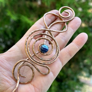 Op de afbeelding: Een haarstick van koperdraad met een gehammerde afwerking en een blauwe agaat edelsteen in het midden. De draad is gedraaid in een spiraalvorm.