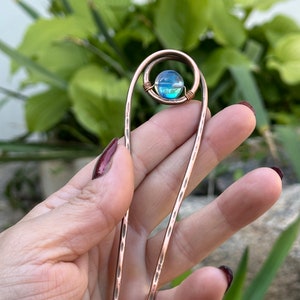 May include: A copper hair stick with a swirl design at the top. The swirl holds a round iridescent blue gemstone.