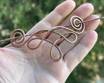 May include: A copper wire hair stick with a decorative swirl design. The stick is held in a hand.