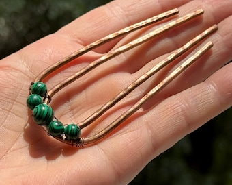May include: Copper hair fork with four tines and a hammered texture. The hair accessory features a cluster of green and black malachite beads. The hair fork is held in a hand against a blurred green background.