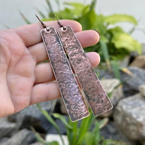 Könnte beinhalten: Ein Paar lange, gehämmerte Kupferohrringe mit rustikalem Finish. Die Ohrringe sind dunkelkupferfarben mit einem leicht rosafarbenen Farbton.