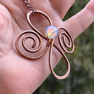 Könnte beinhalten: Kupferfarbene Anhänger-Halskette mit floralem Design und einer irisierenden, runden Perle in der Mitte. Der Anhänger hat eine Kette und wird in einer Hand gehalten. Ein einzigartiges Schmuckstück.