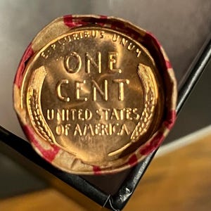 May include: A copper-colored US penny with the words "One Cent" and "United States of America" inscribed on it. The coin is in a red and white paper wrapper.