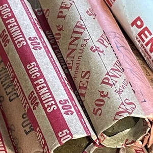 May include: Close-up of several rolls of pennies, each wrapped in paper with red and white stripes. The wrappers are labeled "50¢ Pennies" and "Printed in U.S.A.". The ends of the rolls show the copper-colored coins.