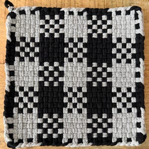 May include: A square potholder featuring a woven checkerboard pattern in black and light gray. The textile construction consists of interlocking loops of yarn, creating a grid design with a small loop at the top corner for hanging purposes.