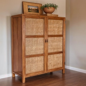 May include: A wooden cabinet with two doors, featuring woven panels and a natural finish. A framed landscape painting and a wooden bowl with greenery sit on top. The cabinet is on a hardwood floor.